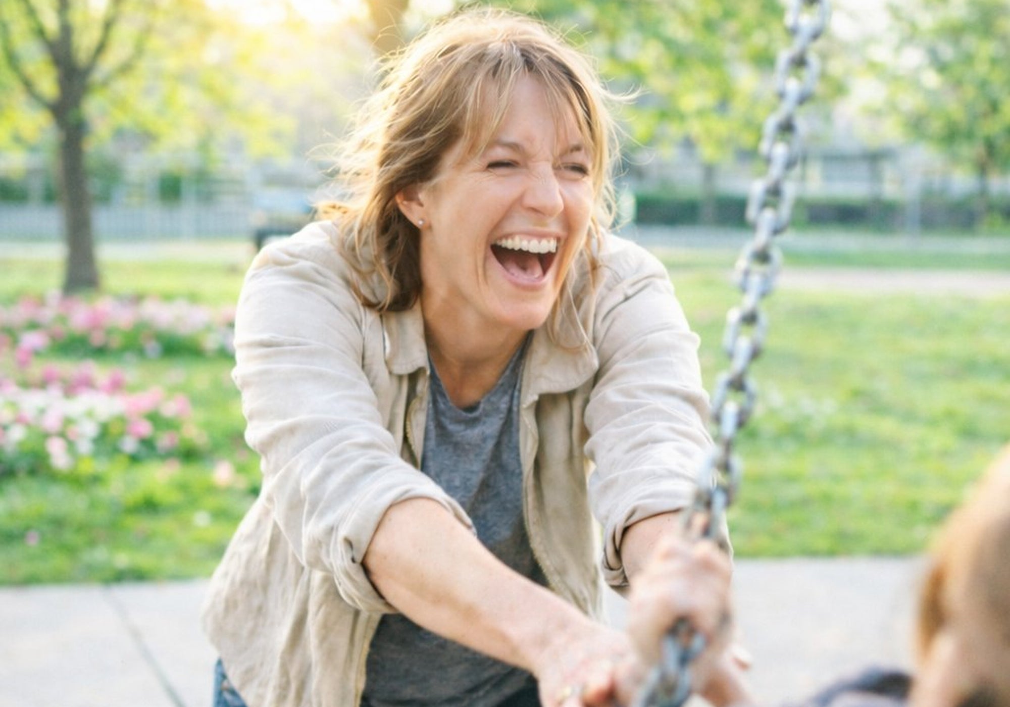 Woman laughing and pushing a child on a swing in a park - gardewear